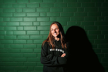 Chiara D´Angelo trägt einen grünen Werder-Hoodie, verschränkt die Arme und steht vor einer dunkelgrünen Wand.