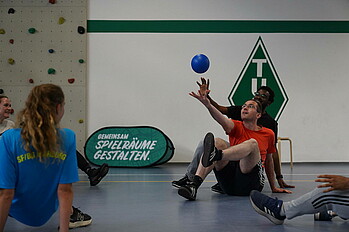 es werden Menschen beim Sport treiben in einer Halle gezeigt. Die Menschen bewegen sich hockend auf dem Boden. rechts im Bild versuchen zwei Menschen in einer sitzenden Position zum Ball zu gehen.