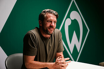 Peter Niemeyer sitzt an einem Tisch, im Hintergrund die Werder-Raute. 