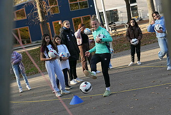 In der Mitte des Bildes ist Lina Hausicke,welche gegen einen Fußball tritt. Im Hintergund stehen sieben Mädchen welche in Rchtung Lina schauen.