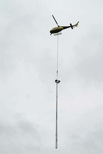 Ein Hubschrauber kreist in der Luft und zieht unter sich einen 20 Meter hohen Flutlichtmasten mit sich.