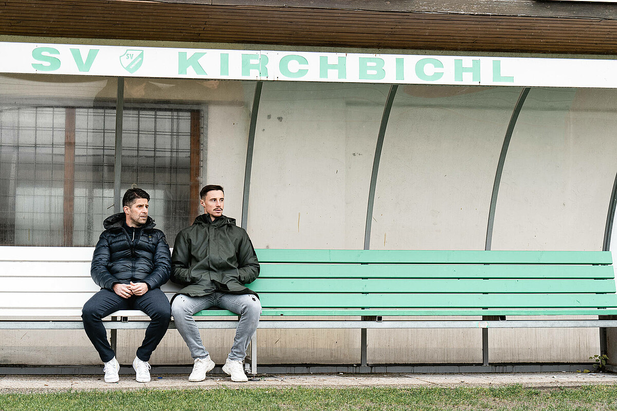 Hanel und Friedl sitzen nebeneinander auf der grün-weißen Bank des SV Kirchbichl. 