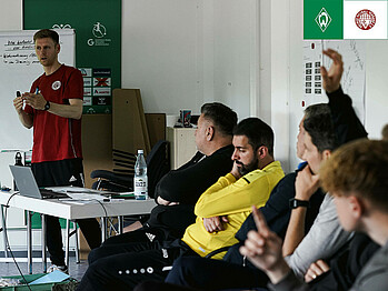 Auf dem Bild sind mehrere Fußballtrainer zu sehen, die in einem Raum sitzen. Ein Referent des Bremer FV gestikuliert mit den Händen, was zeigt, dass er gerade etwas erklärt.