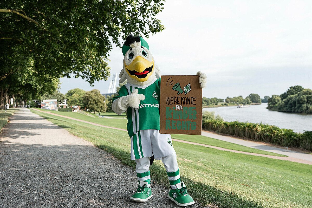 Möwe Toni hält ein Schild mit der Aufschrift Klare Kante für Kinderrechte. Er steht am Deich, im Hintergrund ist das Weserstadion.