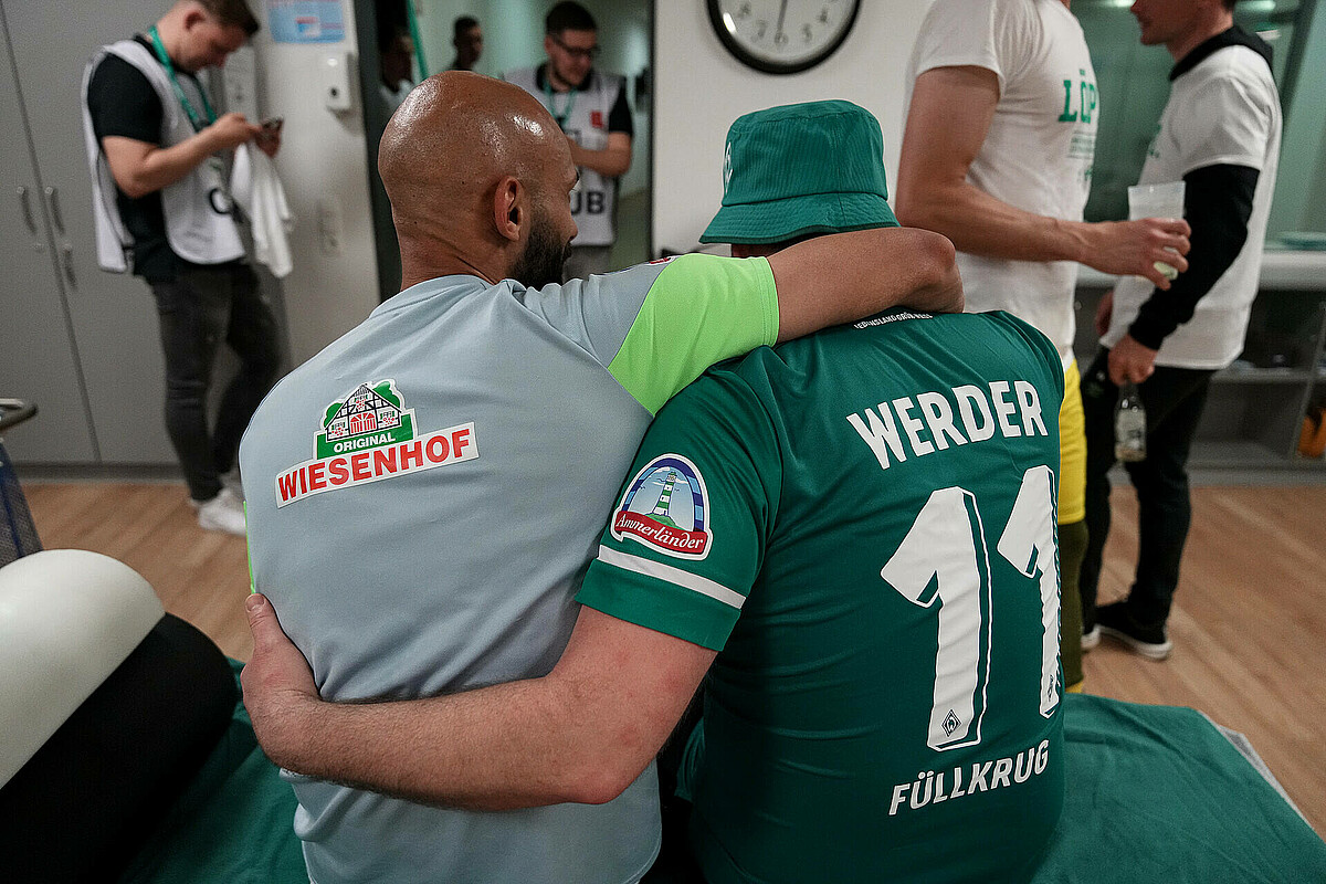 Ömer Toprak und Niklas Füllkrug sitzen Arm in Arm in der Kabine des SV Werder, mit dem Rücken zur Kamera.