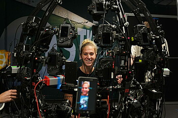 Saskia Matheis lächelt während sie von einem halbrunden Gestell umgeben ist, an dem zahlreiche Fotokameras hängen.