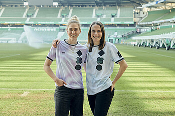 Spielerinnen Saskia Matheis und Reena Wichmann stehen Arm in Arm im wohninvest Weserstadion und lächeln. Sie tragen beide ein Trikot welches von der Organisation COMMON GOAL ist.