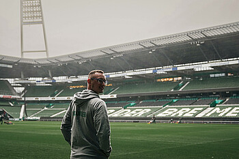 Fabio Ahrens im wohninvest Weserstadion vor dem Spielfeld. Er steht mit dem Rücken zur Kamera und guckt über die Schulter. Auf der Jacke die er trägt steht hin Volunteer drauf.