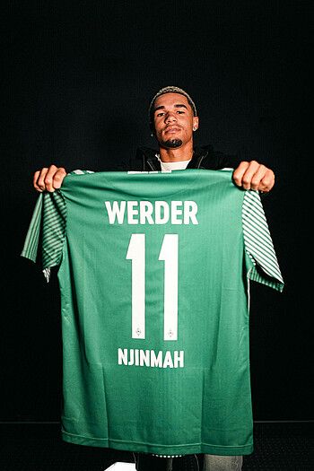 Justin Njinmah hält ein grünes Trikot mit der Nummer 11 und seinem Namen in die Kamera.