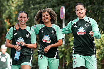Chiara D'Angelo, Amira Arfaoui und Mariella El Sherif tragen grüne Trainingskleidung und lächeln in die Kamera.