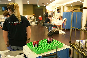 Blutspendeaktion im wohninvest WESERSTADION, ein Tisch mit Blutampullen, links sitzt eine Frau auf deren Poloshirt "schenke Leben, spende Blut" steht.