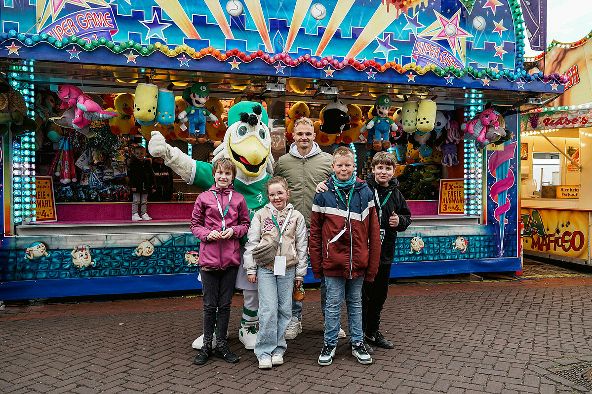 Möwe Toni, Werder-Profi Amos Pieper und Kinder des Kids-Clubs stehen vor einer Bude auf der Osterwiese