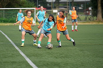 Zwei Spielerinnen in orangen Leibchen versuchen der Spielerin im blauen Leibchen den Ball abzunehmen.