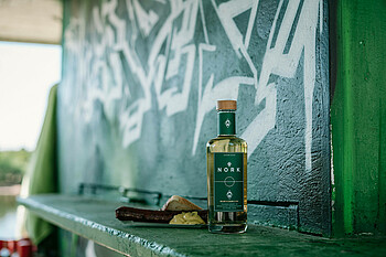 Die Flasche Kurvenkorn von Nork mit einer Bratwurst dahinter vor einer grün-weißen Wand.