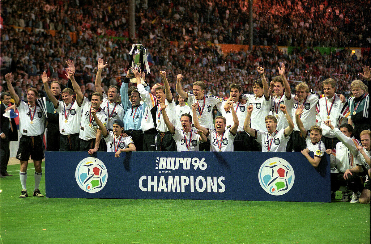 Das Siegerfoto der deutschen Nationalmannschaft 1996, die Spieler jubeln mit der Trophäe hinter einer Bande. 