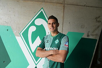 Rafael Borré trägt das Heimtrikot und steht mit verschränkten Armen vor einer gemalten Werder-Raute. 