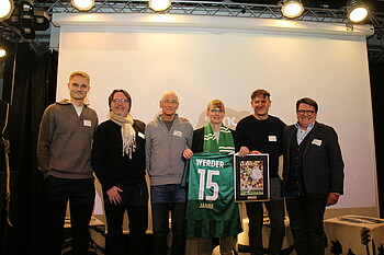 Werder-Spieler Amos Pieper ganz links und Werder-Präsident Hubertus Hess-Grunewald ganz rechts bei der Trikotübergabe für das Lernzentrum Ostkurvensaal