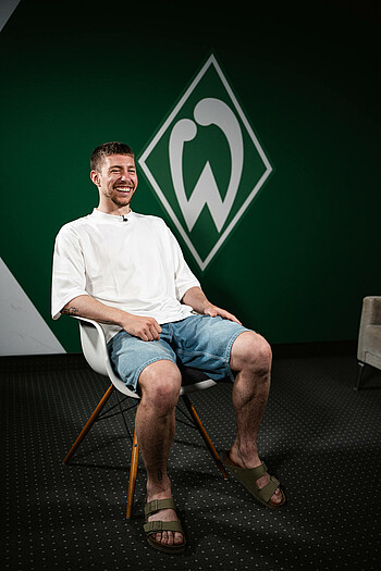 Mitchell weiser lachend im Interview. Er sitzt auf einem weißen Stuhl. Er trägt ein weißes Tshirt, Jeansshorts und Latschen.