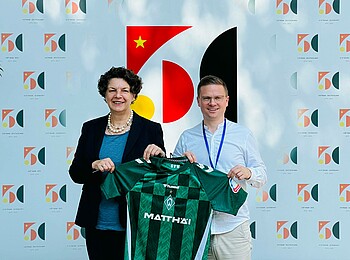 Botschafterin Helga Margarete Barth steht zusammen mit Dominik Kupilas vom SV Werder vor einer Wand. Sie halten ein Werder-Trikot hoch.