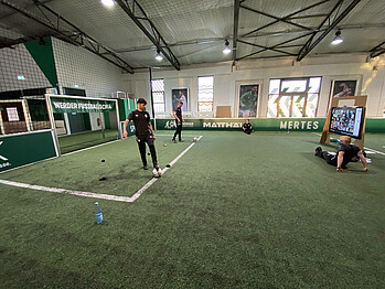 Zwei junge Mitarbeiter der Werder Fußballschule zeigen vor einem Bildschirm Übungen am Ball.