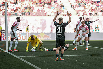 Der Leipziger Keeper hat den Ball am Boden, Romano Schmid ist von hinten zu sehen. Er hebt beide Arme.