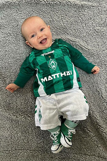 Baby Keke Massalme im Werder-Trikot, liegt auf dem Rücken auf einer Decke und lacht in die Kamera. 