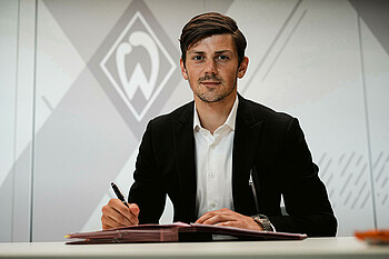 Dawid Kownacki sitzt mit Sakko am Tisch und unterschreibt seinen Vertrag. Im Hintergrund eine graue Werder-Raute.