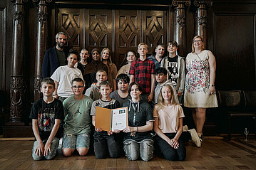 Zusehen ist ein Gruppenfoto. Die Klasse der Gaußschule zwei Bremerhaven steht mit ihrer Klassenlehrerin und Henrik Oesau vor einem großen Türrahmen. Sie befinden sich im Bremer Rathaus. 