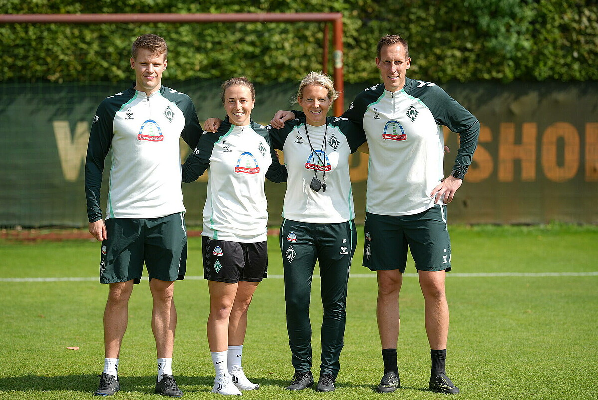 Vier Personen stehen in weißen Sportshirts und dunkelgrünen Hosen Arm in Arm auf einem Fußballfeld.
